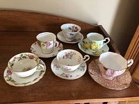 Photo showing six assorted vintage tea cups and saucers with floral patterns and gold trim on wooden surface.