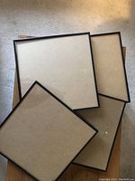 Five black rimmed frames stacked and laid on carpet floor showing back side with fastening hardware on each.