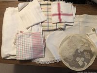 Various antique linens including white cloths with lace, checked fabrics, and embroidered doilies arranged on a wooden surface.
