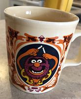 Close-up photo of a ceramic mug featuring a vintage-style Muppet character named Animal surrounded by orange and purple decorative frame.