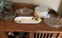 Photo of six items in the lot displayed on a wooden cabinet including two casserole dishes with lids, two glass bowls, one ceramic decorative plate with fruit design, and one cut glass serving bowl.