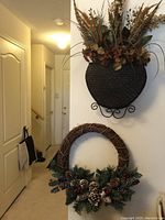 Two seasonal door wreath hangings displayed on a wall in a hallway. One is a grapevine wreath with pine cones and greenery; the other is a dried flower arrangement in a black textured metal holder with stand.