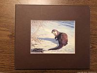 Mounted print showing a realistic otter on snow near water with trees in background