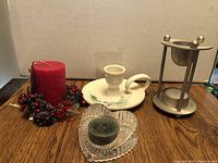 Four items on a wooden surface: small red scented candle with berry wreath, white pottery candle holder, clear heart-shaped glass holder with green candle, and metal incense burner
