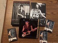 Photo of three Bruce Springsteen books and three cassette tapes arranged on wooden surface, showing covers and front of items
