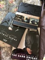 Three hardcover books laid out on a floral quilt with Batman artwork on the covers; titles include 'Batman Begins,' 'The Dark Knight Manual,' and 'The Art and Making of The Dark Knight Trilogy'