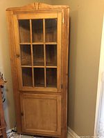 Front view of the vintage wooden corner cabinet, showing the closed upper glass door with wooden grid panels and closed lower solid wooden door.