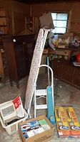 Photo showing folding ironing board with cover and iron holder, vintage vacuum, white step ladder, and assorted household items in basement.