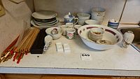 Wide shot showing entire collection including carved wooden chopsticks with red tips, multiple stacks of ceramic bowls, small floral cups and spoons on a kitchen counter.