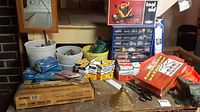 View of the workbench with various boxed nails, bench vise, plastic buckets, and blue multi-drawer hardware organizer, along with toolbox and hand tools scattered on the table.