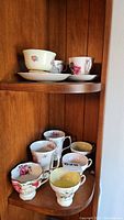Photo of 8 assorted china teacups and saucers with floral patterns on wooden shelves, showing variety and condition including chips