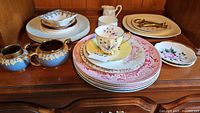 Wide view of various china plates, teacup, and brass-accented blue creamer set on wooden surface