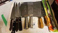 Photo shows 8 cutting tools including knives and cleavers arranged on a white cloth, highlighting wooden and black handles and metal blades with varying sizes and types.