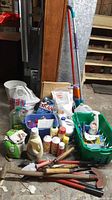 Full view of the lot showing household cleaners, mops, a bucket, and garden tools organized in a basement setting.