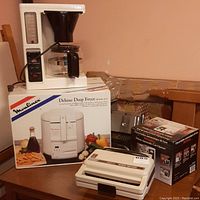 Full lot showing Melitta coffee maker, Moulinex Deluxe Deep Fryer in box, Sunbeam Snack Factory sandwich maker, and automatic can opener in box on wooden table.