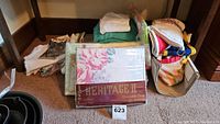 Lot overview showing folded linens on carpet including pack of Heritage II twin bed sheets, towels, tablecloths, aprons and oven mitts.