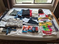 Wide shot showing the laid out men's clothing items including folded pants, sweaters, vests, T-shirts, and small packaged handkerchiefs on a white bed sheet under a window.