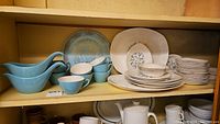 Shelf with blue gravy boats, blue cups, white bowls, and patterned plates and trays.