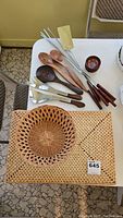Photo showing wooden serving utensils, metal barbecue skewers, small wooden bowls on a table with two rectangular woven placemats beneath them.