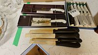 Photo of Royal Albert cheese cutter knives with white handles, two knives in original boxes, and a set of six steak knives with black handles placed on a countertop.