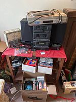 Full view of stereo system and audio/video items on table, including stereo components, headphones, VHS rewinder, and boxes of DVDs and VHS tapes on floor.