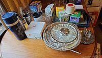 Wide shot showing collection of thermos, travel mugs, glass trays, boxed mugs, tea boxes, and other kitchen accessories on wooden table.