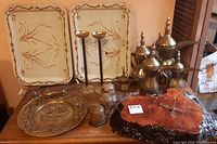 Front view showing metal bed trays in background, five brass tea servers grouped, hanging bells, brass wall plate, and wooden clock with natural wood finish