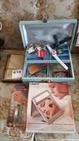 Photo showing light blue two-tier jewelry box open with multiple watches inside including X-Files and Calgary Olympics themed watches, silver change purse, Clairol lighted mirror box, and wrist blood pressure monitor.