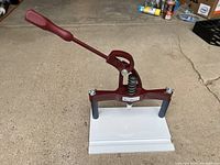 Full view of the BenchMaster picture frame joining press showing red metal lever handle, base, and rubber feet on the ground.