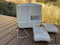 Beige/white large humidifier shown from side view on outdoor wooden deck, with two sealed replacement wick filters next to it.