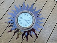 Bulova sun wall clock lying on wooden decking showing the metal sunburst frame and round white clock face with Arabic numerals.