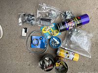 Photo showing all items in the lot laid out on carpet. Items include metal wire cable, multiple small bags with screws and hooks, corner protectors, tape rolls, and adhesive spray cans.