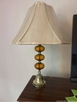 Vintage table lamp with amber glass globes stacked on a brass base and a cream fabric lampshade, shown unlit on a wooden surface.