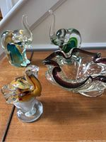 Four art glass items on wooden floor: three elephant paperweights in different colors and one free-form bowl.