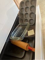 Photo showing a 12-cup metal muffin tin, clear rectangular Pyrex baking dish, wooden rolling pin with red handle, and several metal cookie sheets.