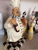 Close-up of the largest chef figurine holding a wine bottle and bread, showing intricate details of face, clothing, and base.