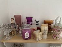 Photo shows assortment of candles, candle holders, vintage figurine, and decorative glass items on a shelf.