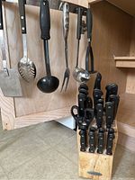 Farberware knife block with multiple knives and kitchen scissors next to hanging kitchen utensils on a metal bar in wooden cabinetry.