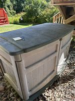 View showing overall rectangular deck box with beige body and greenish lid, closed and outdoors on gravel under a wooden deck.