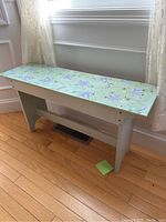 Full view of a mint green decorative wooden bench positioned near a window on wooden floor. Shows the full bench shape and size.