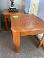 Pair of wooden end tables shown side by side with a lamp on one table. Square tops with rounded edges, visible parquet wood pattern, rounded legs.
