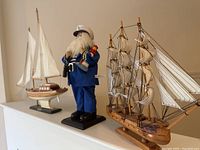 Three items shown together: two wooden model boats and a fishing captain figurine between them, displayed on a white shelf.