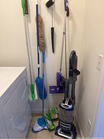 Overall photo showing Shark vacuum alongside duster tools and Swiffer sweeper with refills, placed near laundry area.