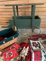 Full view of green wooden holiday cart with attached red wheels and handle, next to woven basket, assorted holiday fabrics and boxes of Christmas cards on the floor.