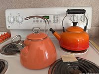 Two kettles on stove; one bright orange Le Creuset and one light orange/peach kettle