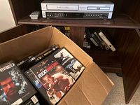 Photo shows a silver Carver 4-head DVD/VHS player on a wooden shelf with some VHS tapes beside it and a remote on top.