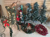 Wide photo of the illuminated deer adorned with lights and red bow, several snow-frosted pine trees, and two holiday wreaths with pinecones and red berries on the ground.
