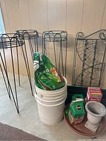Black metal plant stands, white buckets filled with soil amendment bags, and black rectangular planter trays against a beige wall.