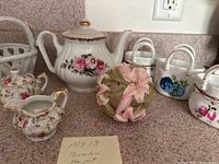 Overall lot including porcelain teapot, creamer, sugar bowl, two porcelain baskets, and decorative floral ball arranged on countertop.
