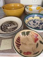 Photo of 13 ceramic bowls including mustard yellow nesting bowls, various decorative pasta bowls with floral and fruit motifs.